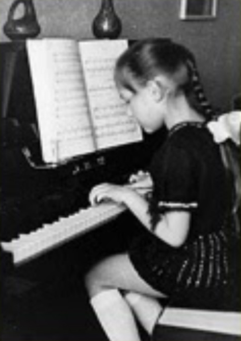 Katya Grineva as a young girl, seated in profile at an upright piano, reading sheet music with her hands on the keys, circa 1977