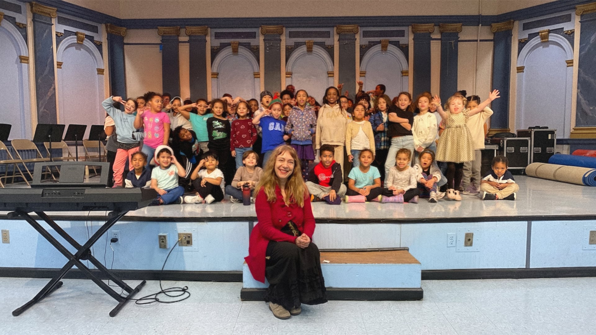 Katya Grineva seated at the edge of a school stage beside a keyboard, surrounded by a large group of joyful children after a concert