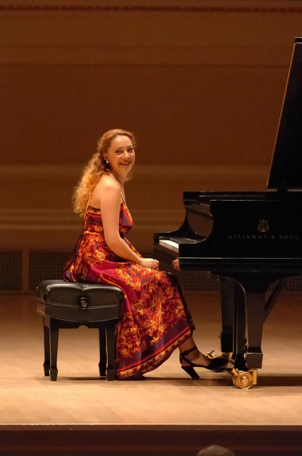 Katya Grineva smiles from the piano bench beside the Steinway on the Carnegie Hall stage, in a deep red gown