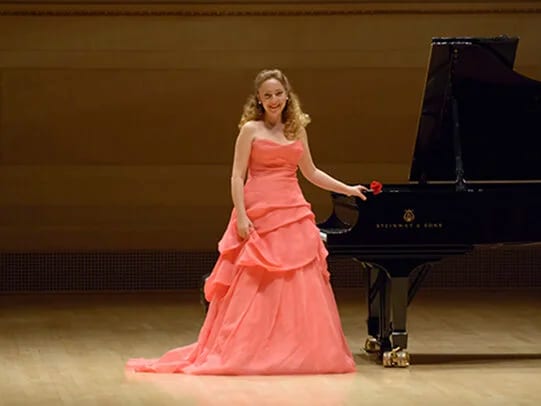Katya Grineva in a flowing coral gown beside the Steinway grand on the Carnegie Hall stage, a single rose in hand