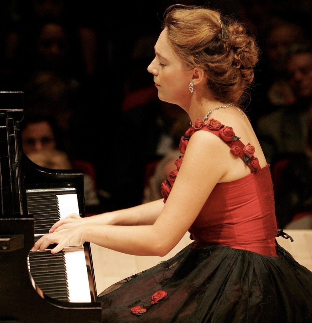 Katya Grineva performing at a grand piano in a deep red gown under stage light