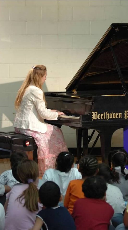Katya Grineva plays piano in a school gymnasium as children sit on the floor watching