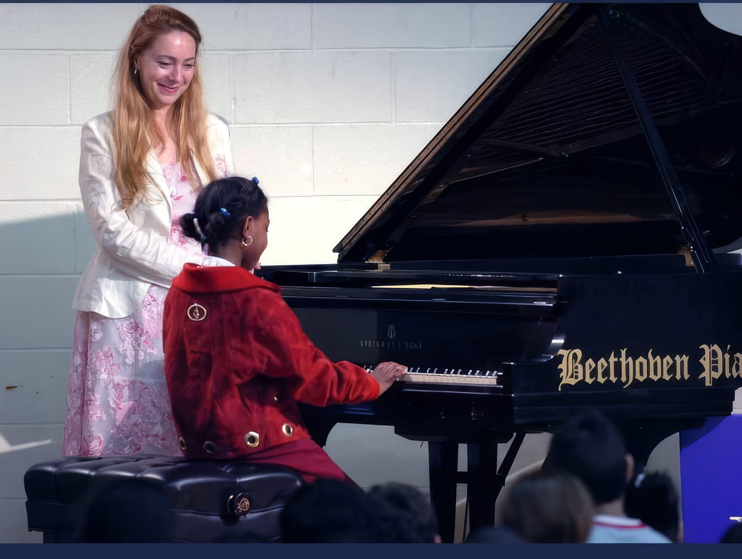 Katya Grineva stands beside a young student playing a grand piano at a school concert